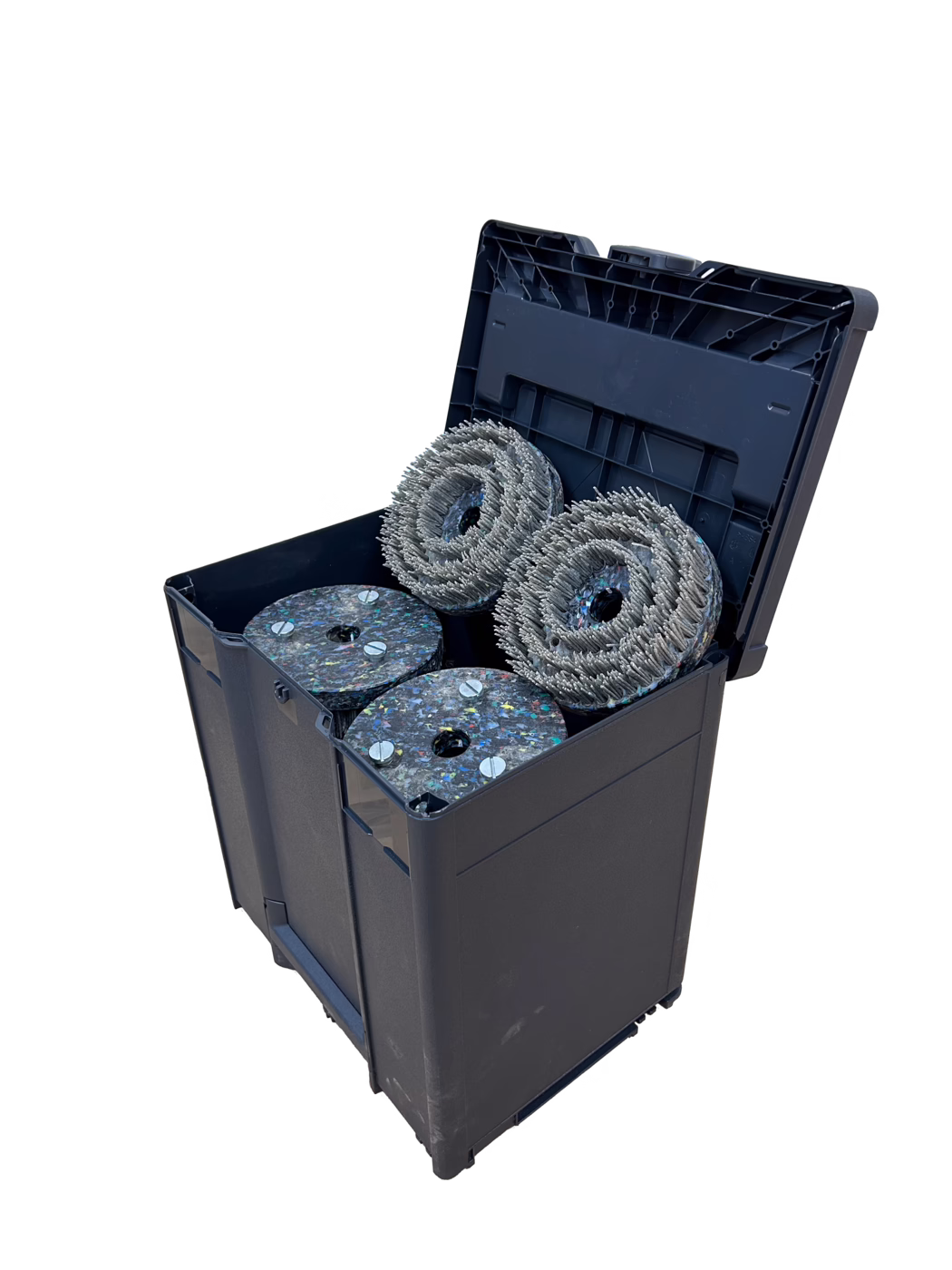 A sturdy black plastic storage container with its hinged lid open, revealing four large circular tool heads inside. Two of the items are dense abrasive brush wheels with metallic bristles wound in concentric rings; the other two are thick composite polishing pads made from multicolored recycled material and secured with visible metal screws. The container shows minor scuffs and signs of use; the photo is taken on a clean white background with soft shadows, emphasizing the texture of the bristles and the recycled pad material. The objects appear to be replacement heads for industrial floor cleaning or surface preparation equipment.