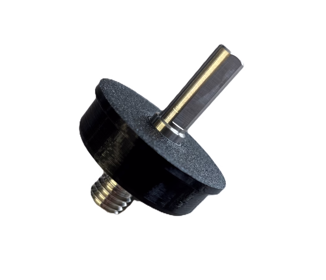 Close-up of a small mechanical assembly: a hexagonal metal shaft with a brass-colored coating inserted into a black plastic or 3D-printed knob or spacer. The metal shaft shows a polished flat face and visible machining marks; a threaded metal stud with concentric screw threads is visible at the base. The black part has a textured, layered surface typical of FDM 3D printing. The object is photographed diagonally on a clean white background.